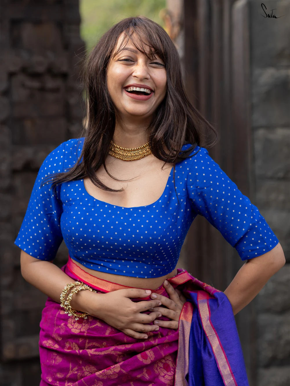 Woman wearing a blue blouse with white polka dots and a pink saree, standing against a dark background.