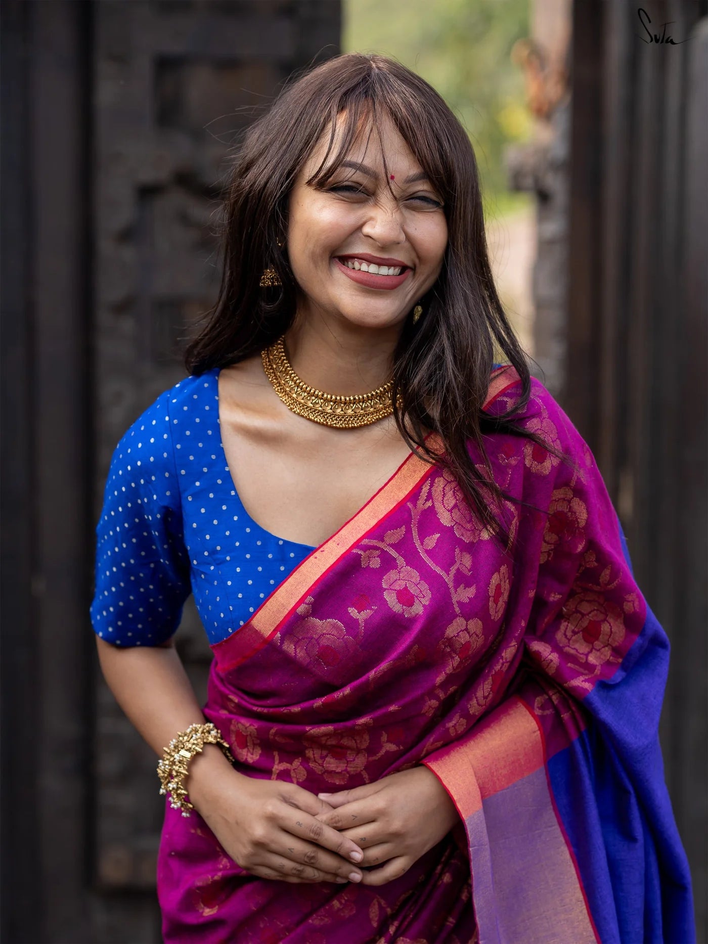 Woman wearing a blue blouse with a purple saree, smiling outdoors.