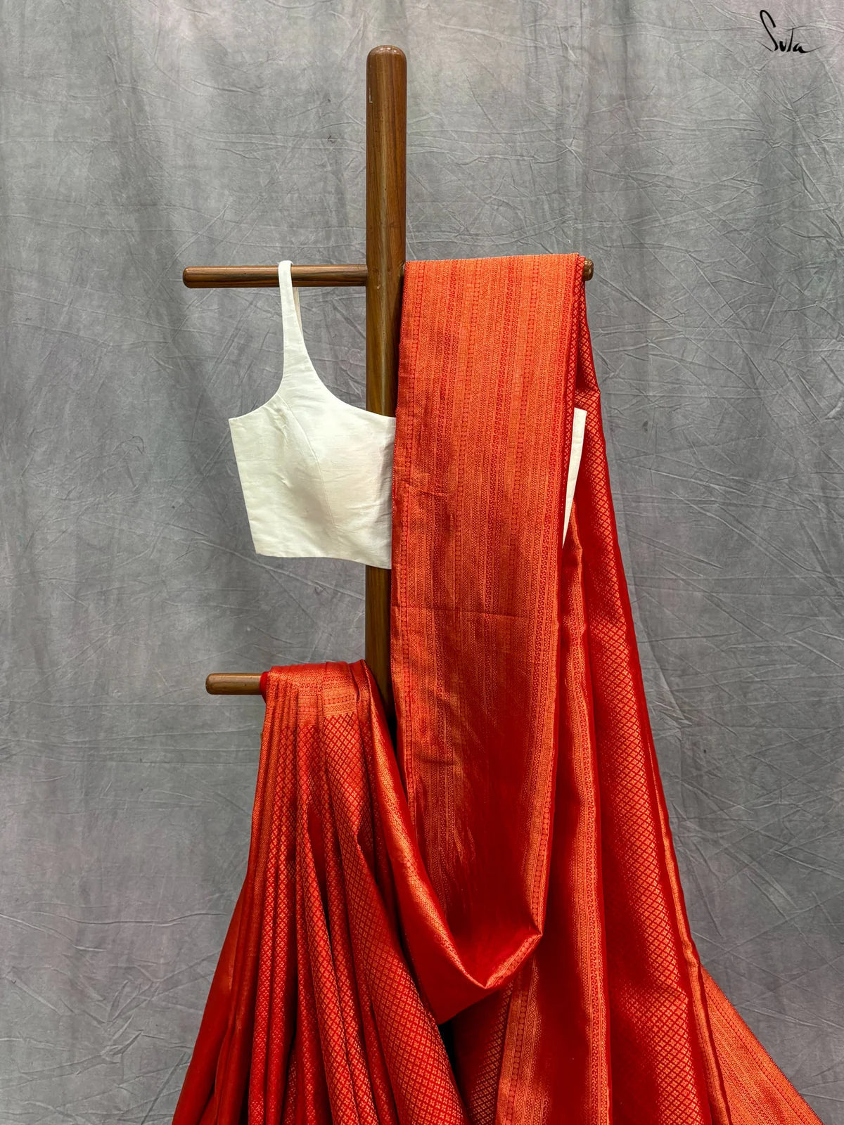 Red saree with gold zari and a white blouse draped over a wooden stand against a gray background.