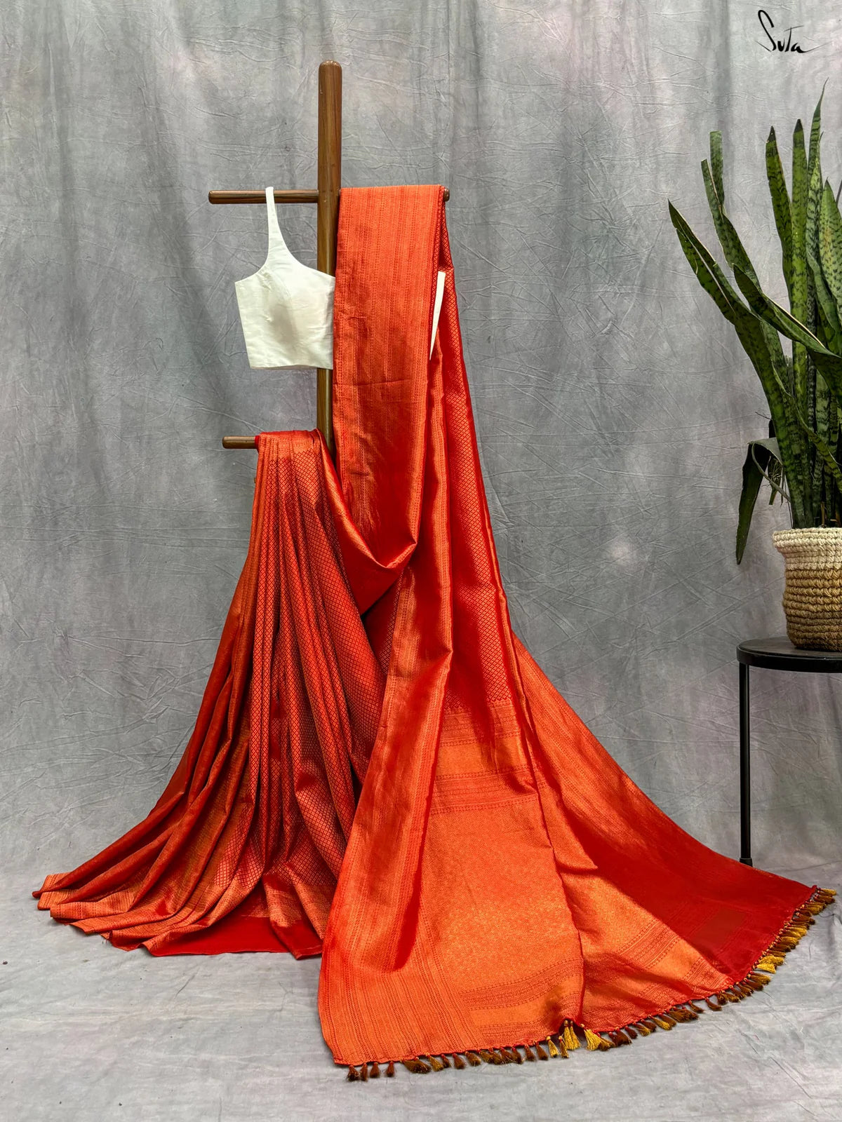 Red saree with gold zari and a white blouse draped over a wooden stand against a gray background.