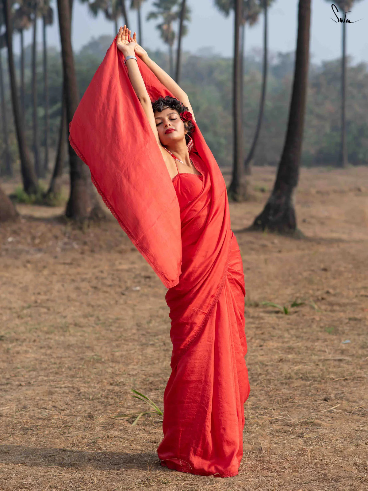 Woman in a red saree holding it up in an outdoor setting with trees in the background