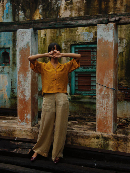 Person wearing a mustard yellow shirt and beige pants standing in front of a dilapidated wall.