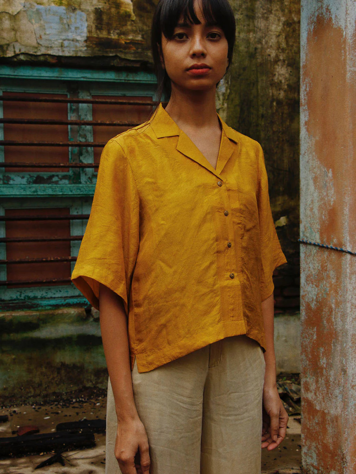 Person wearing a mustard yellow shirt and beige pants standing in front of a textured wall.