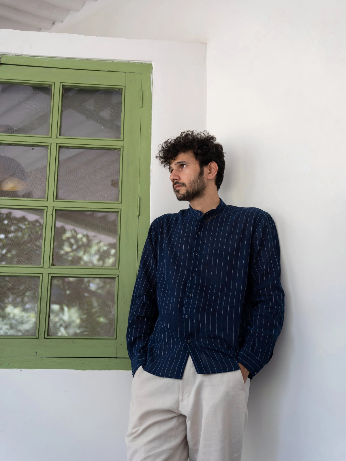 Man wearing a dark blue striped shirt and light-colored pants leaning against a white wall with a green-framed window.