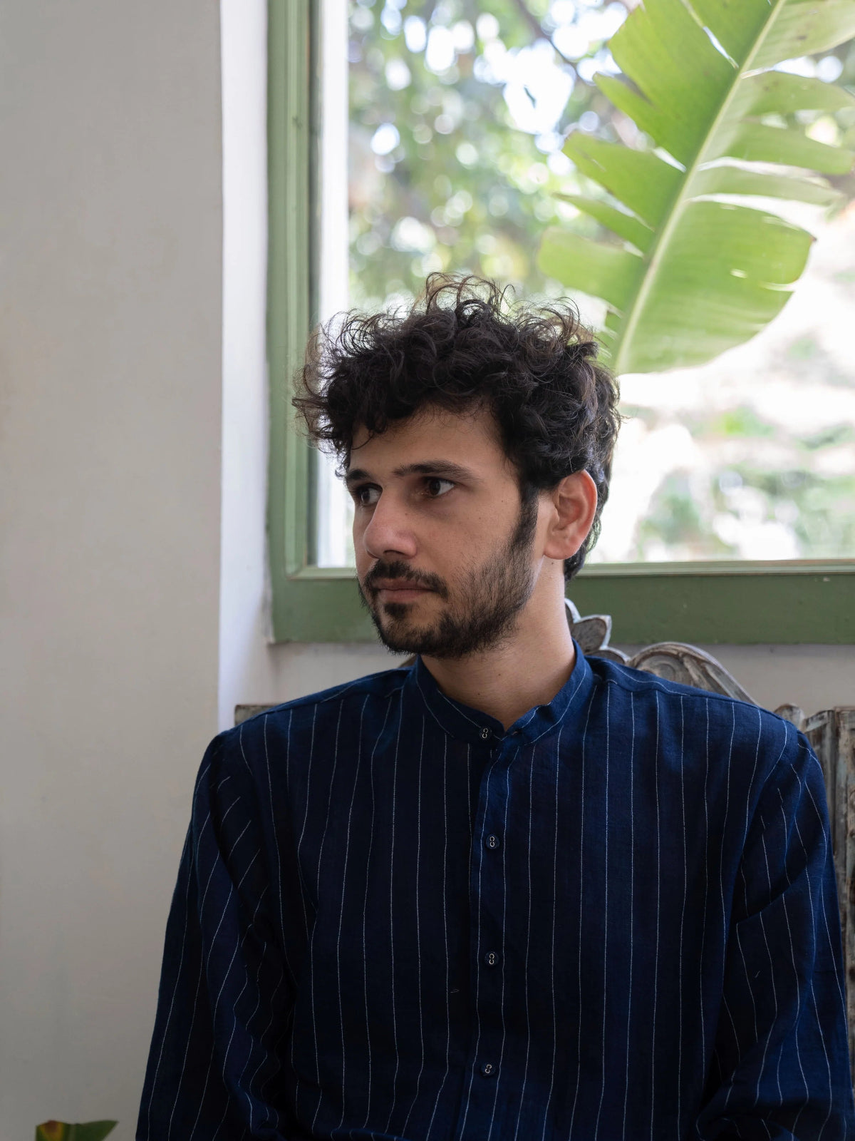 Man with curly hair and beard wearing a blue striped shirt sitting indoors.