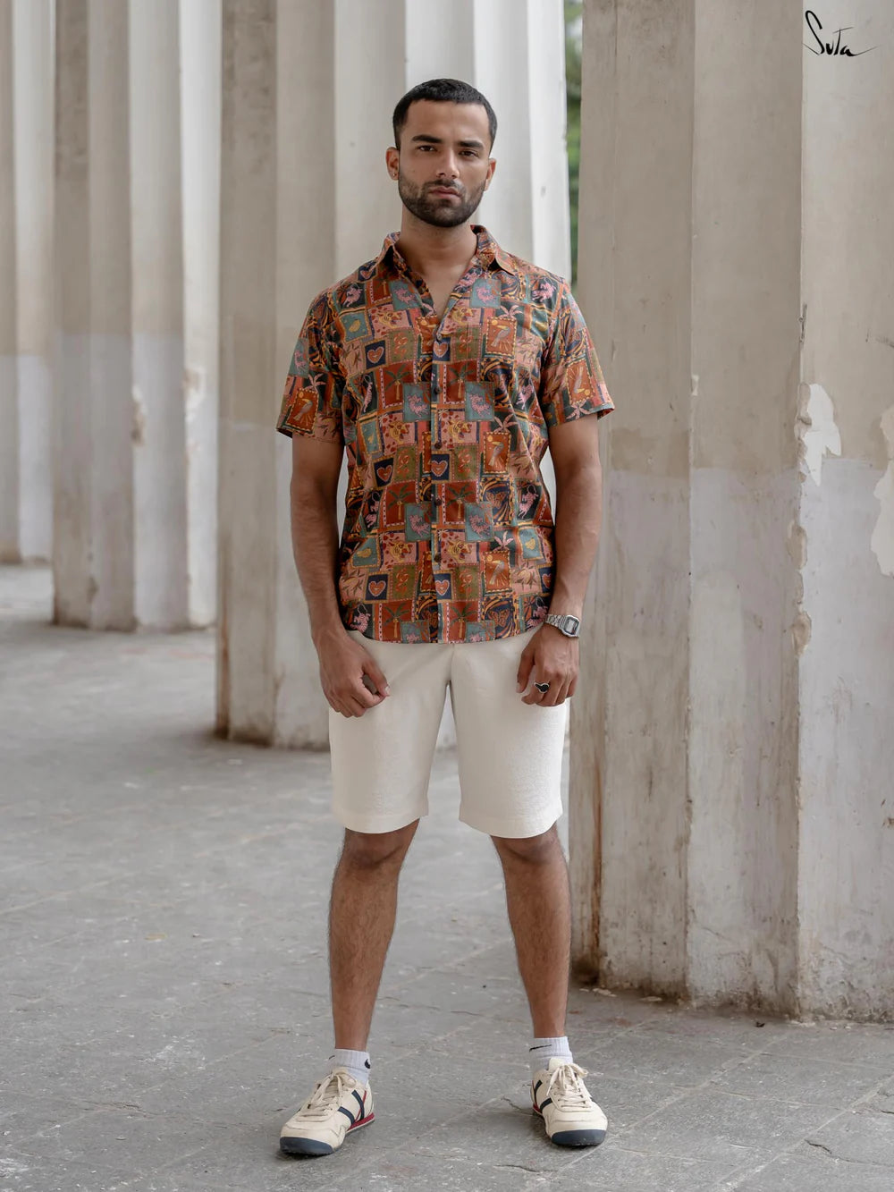 Man wearing a multicolored patterned shirt and beige shorts standing in an outdoor setting with columns.