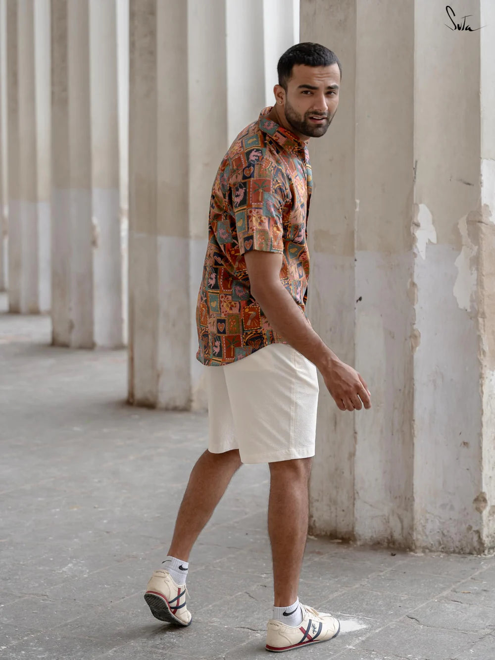 Man wearing a patterned shirt and white shorts standing in an urban setting with columns.