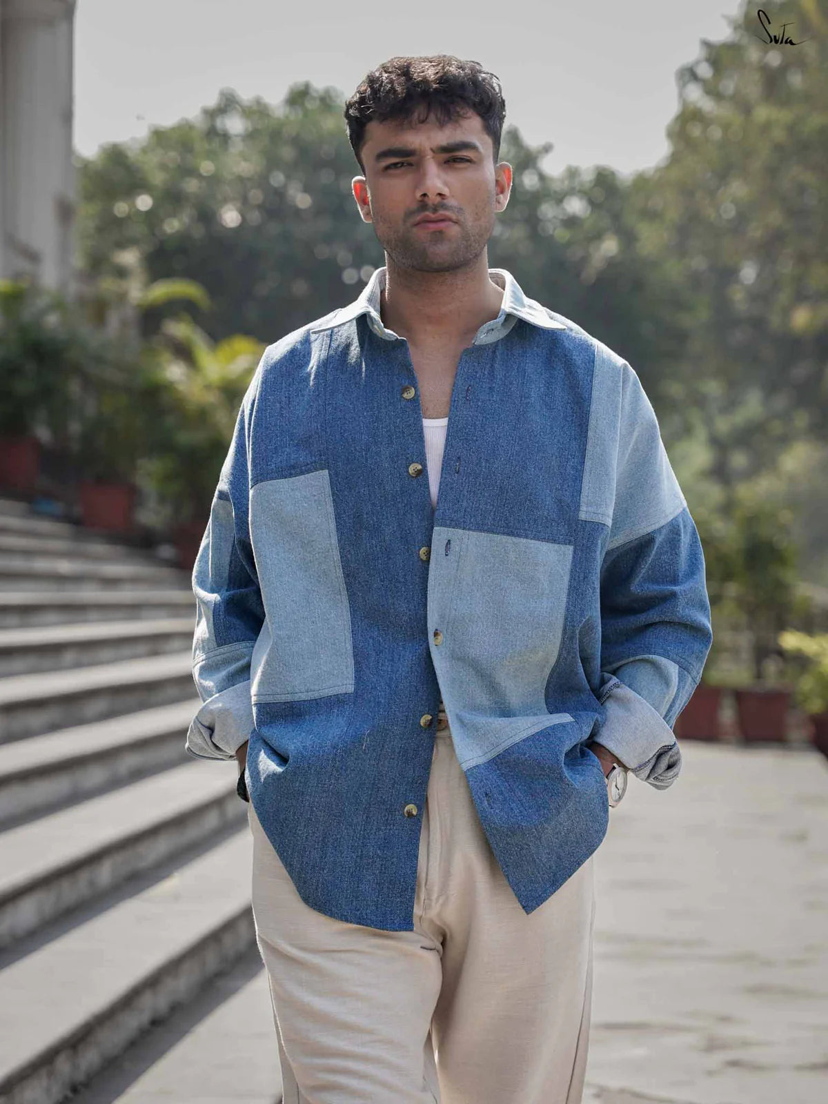 Man wearing a blue patchwork denim shirt standing on steps outdoors with greenery in the background