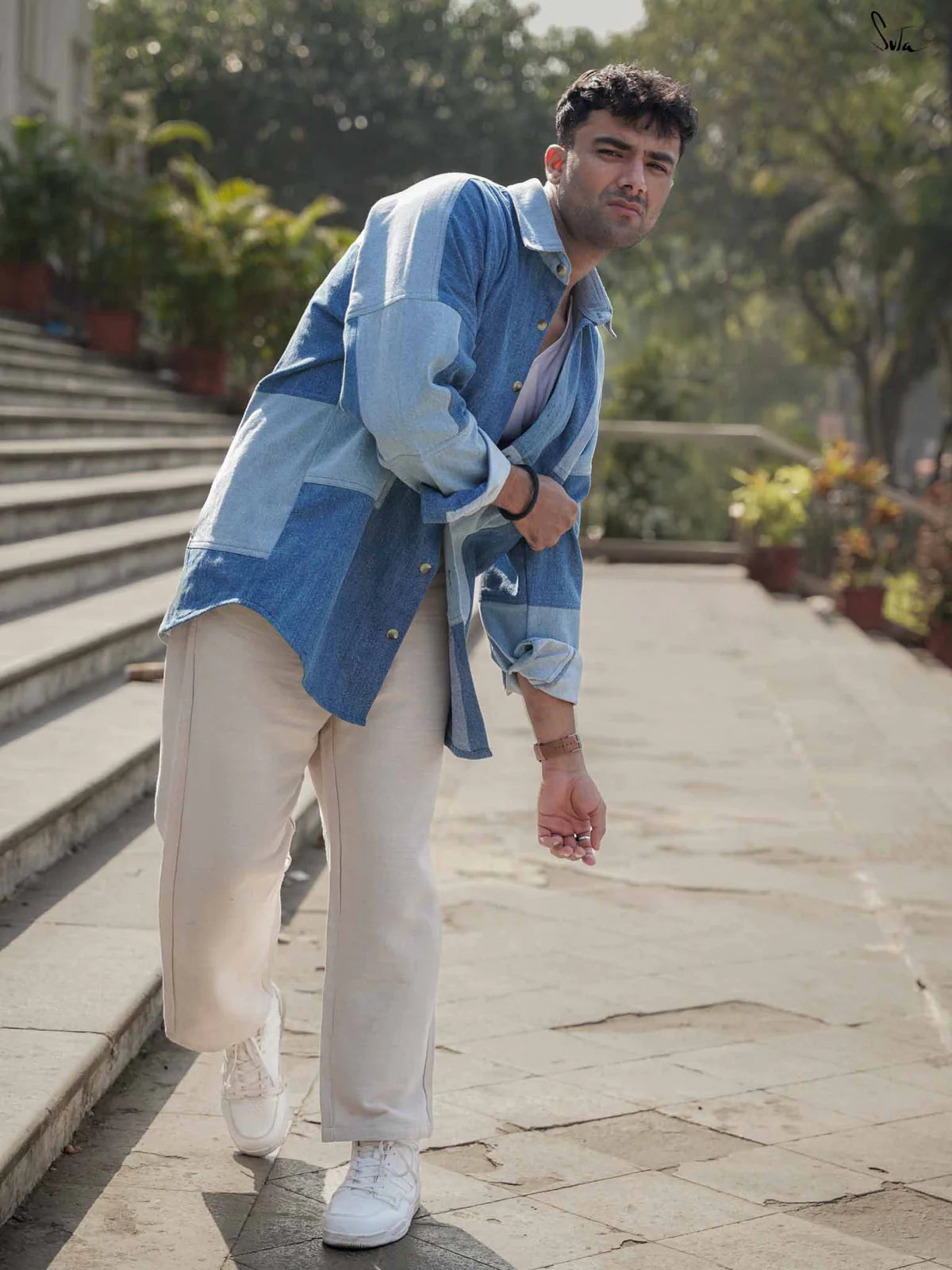 Man wearing a blue denim shirt and beige pants standing on a stone pathway with plants in the background