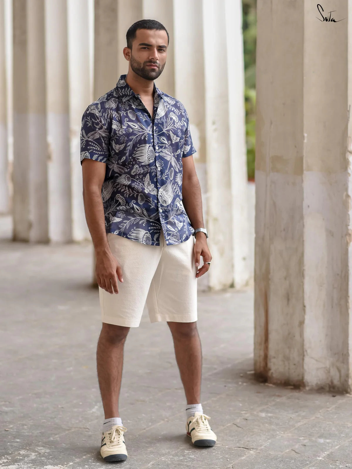 Man wearing a blue patterned shirt and white shorts standing in front of white columns.