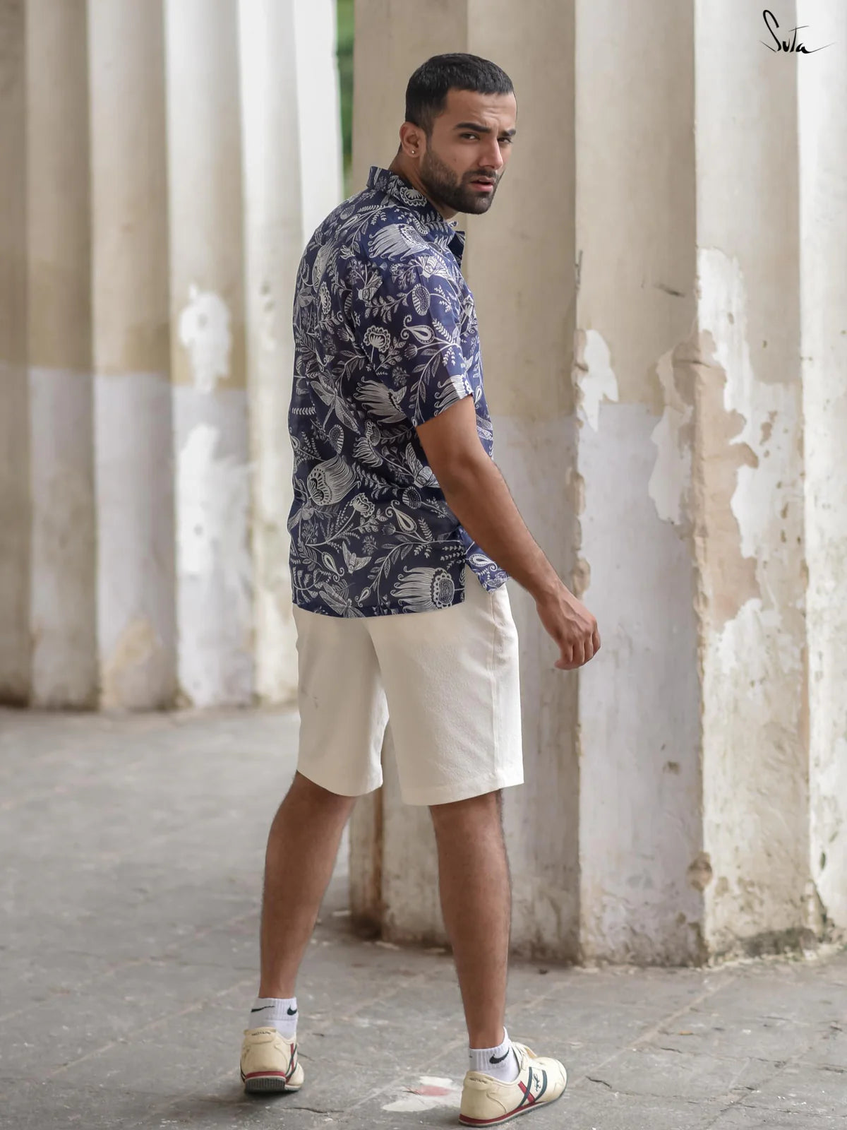 Man wearing a blue patterned shirt and white shorts standing in front of white columns.