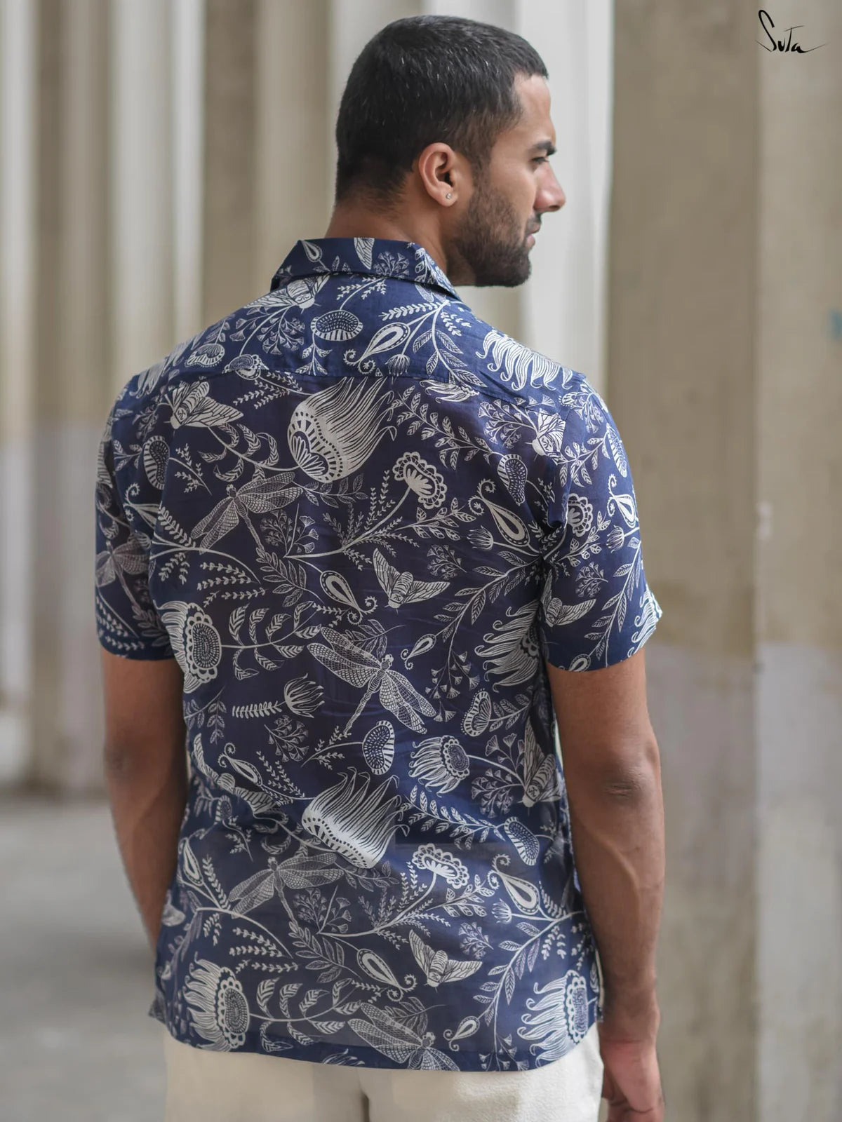 Man wearing a blue patterned shirt and white shorts standing in front of white columns.