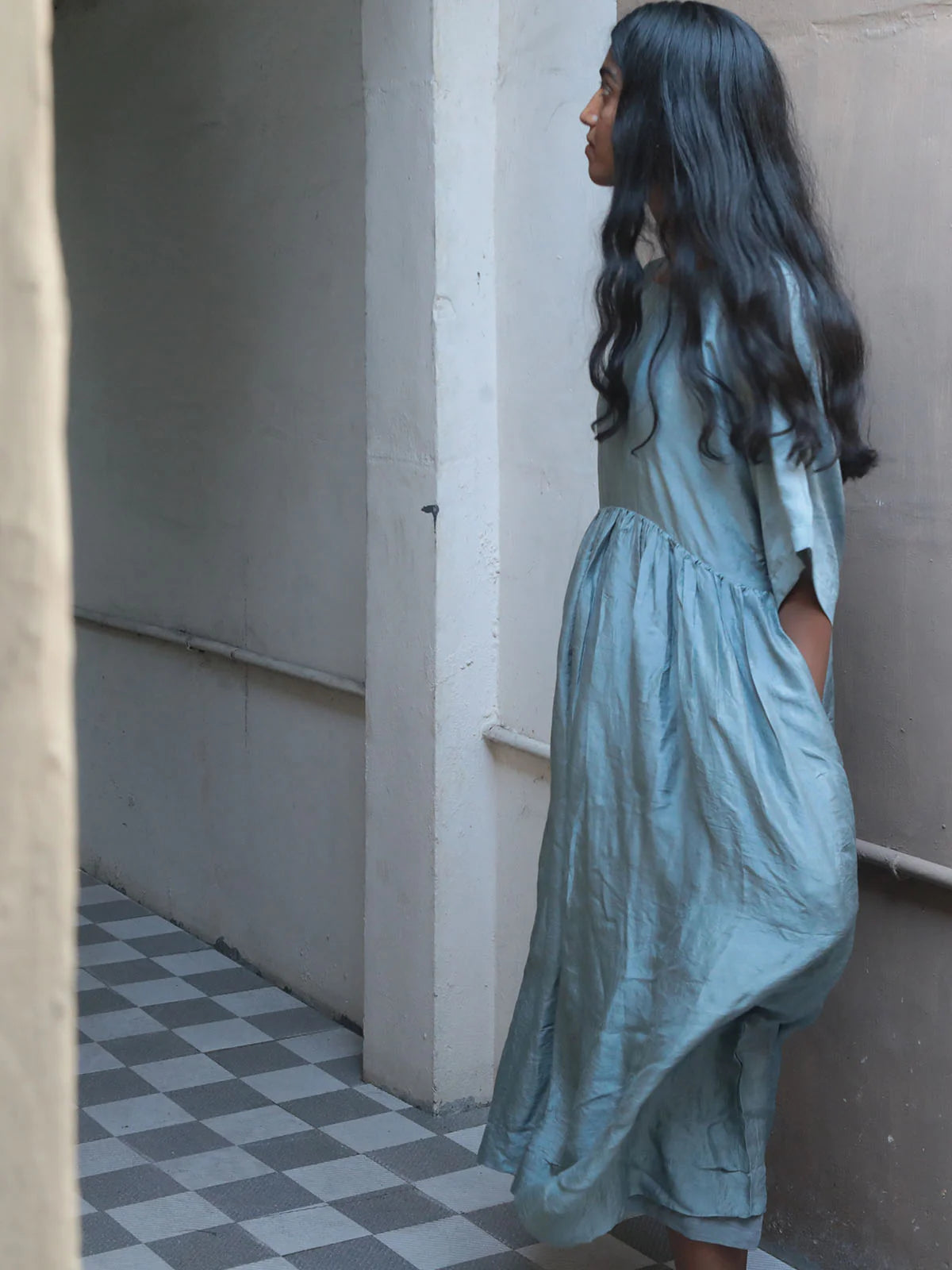 Woman in a blue dress standing in a narrow hallway with checkered floor.