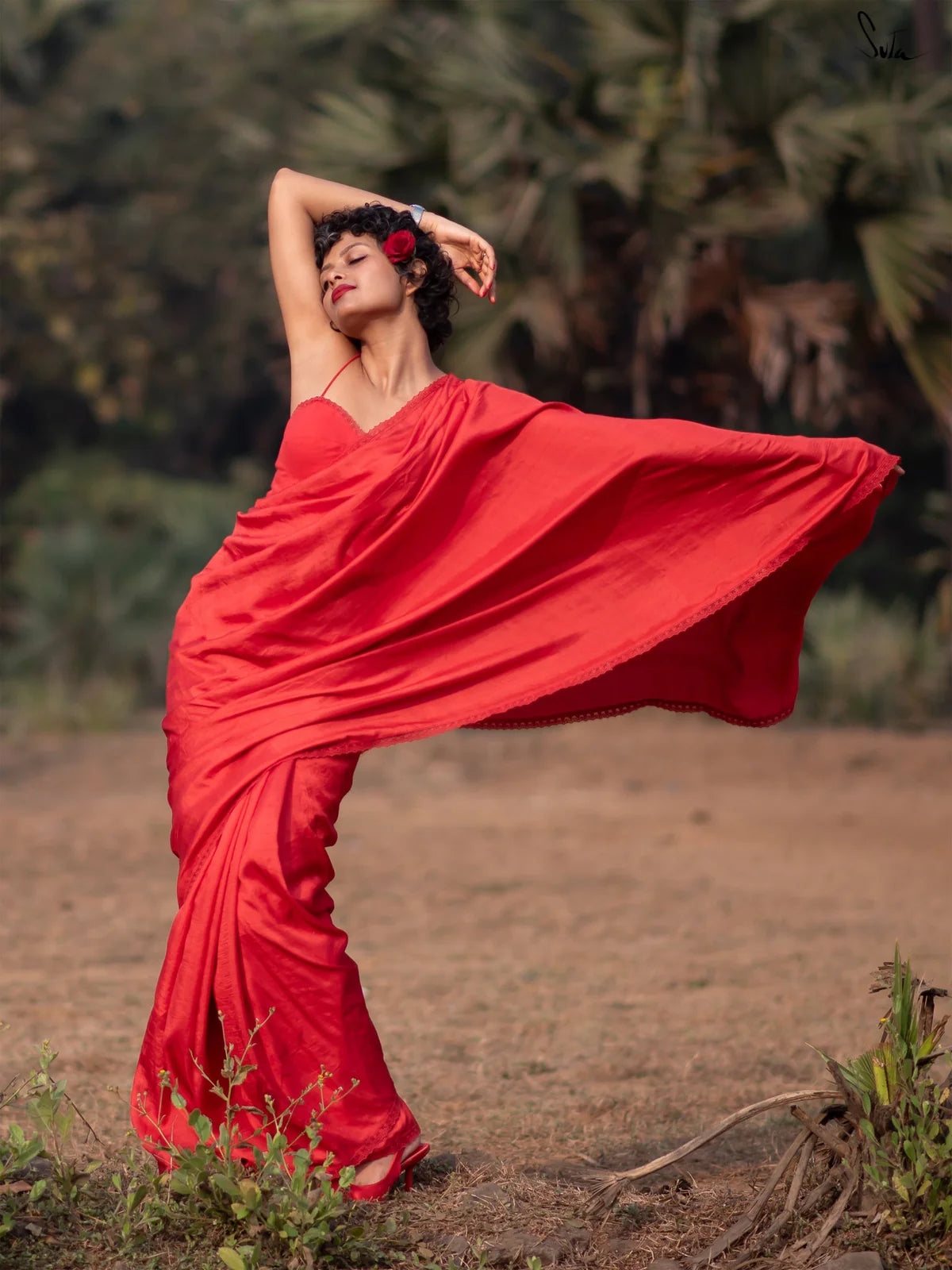 Woman in a red saree dancing outdoors with greenery in the background