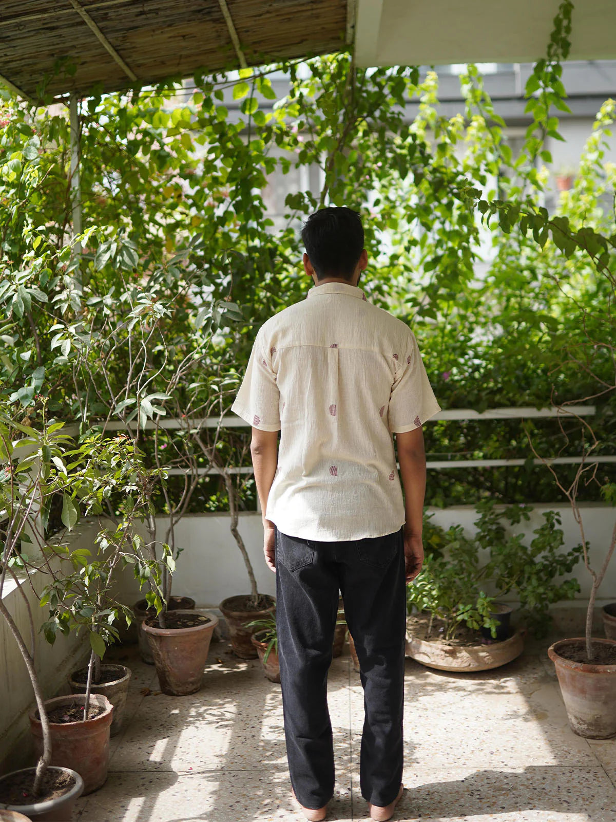Person in beige shirt with half moon red pattern tending to plants on a balcony with buildings in the background