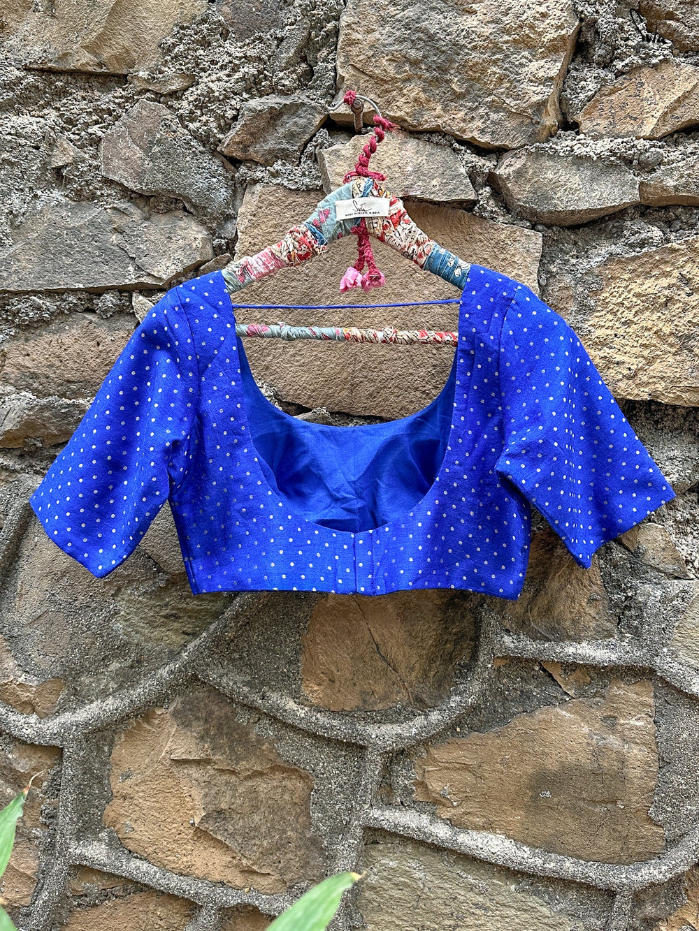 Blue blouse with white polka dots on a floral hanger against a stone wall.