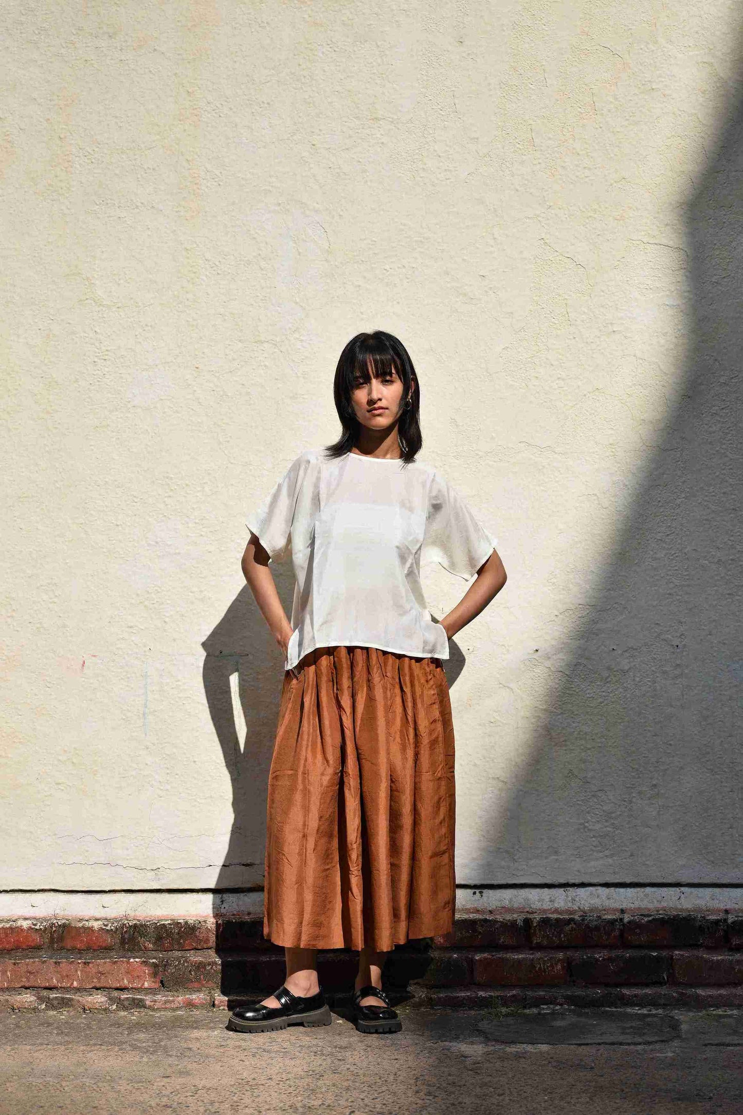 Woman wearing a white top and brown skirt standing against a light-colored wall.