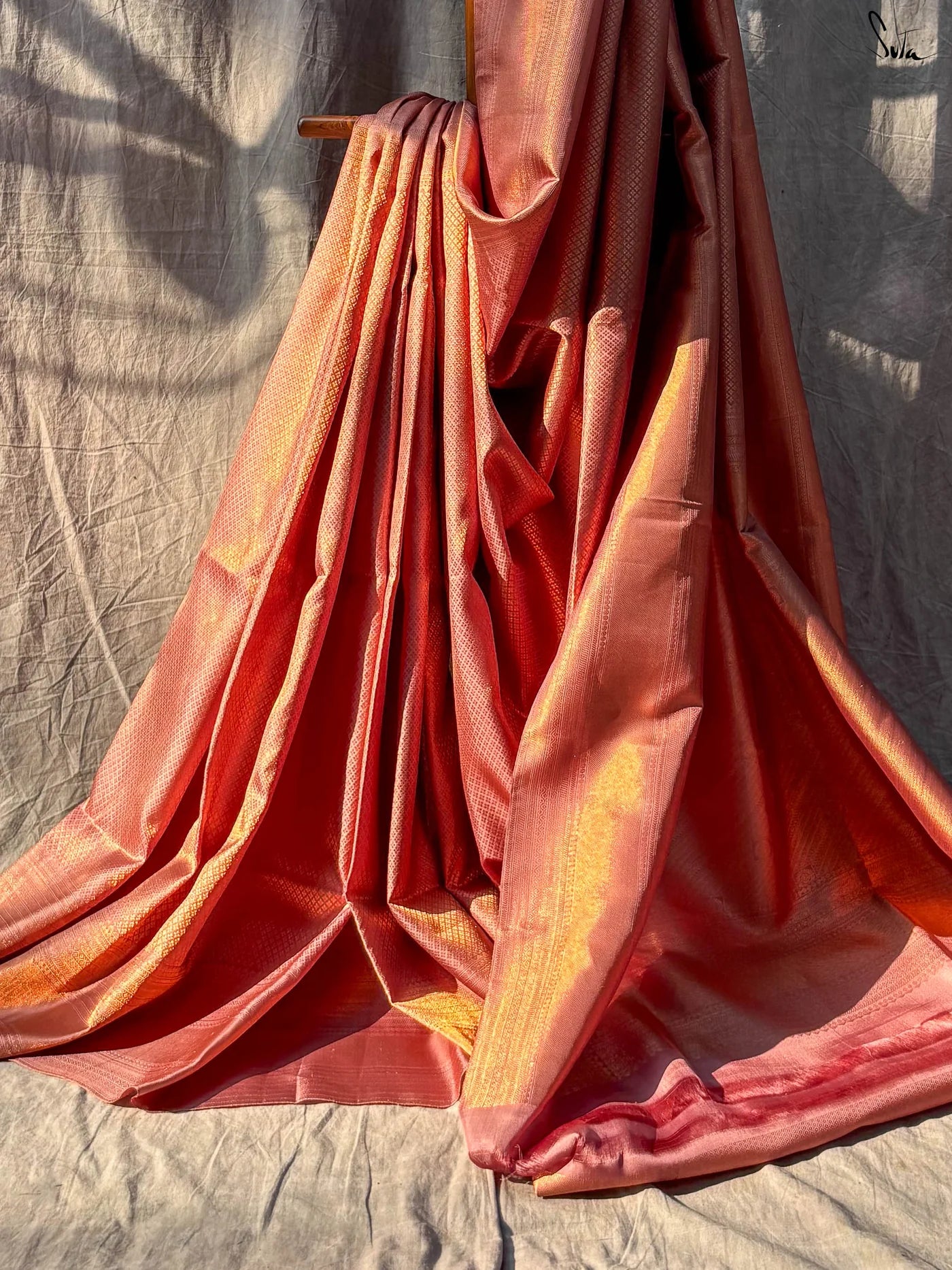 Red and Gold saree fabric draped over a wooden stand against a textured wall.