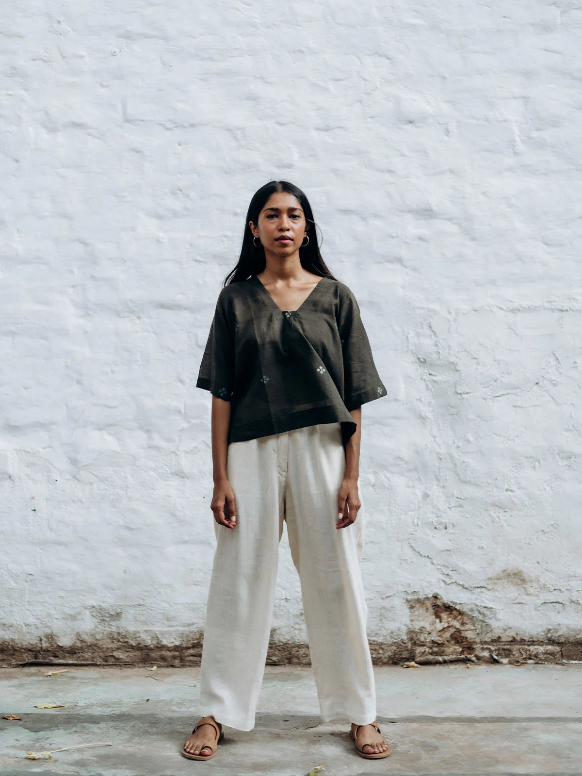 Woman wearing a gray top and white pants against a white brick wall.