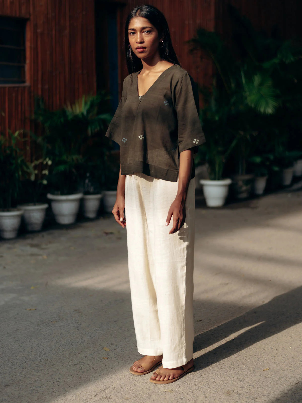 Woman wearing a gray top and white pants against a wooden wall and plants