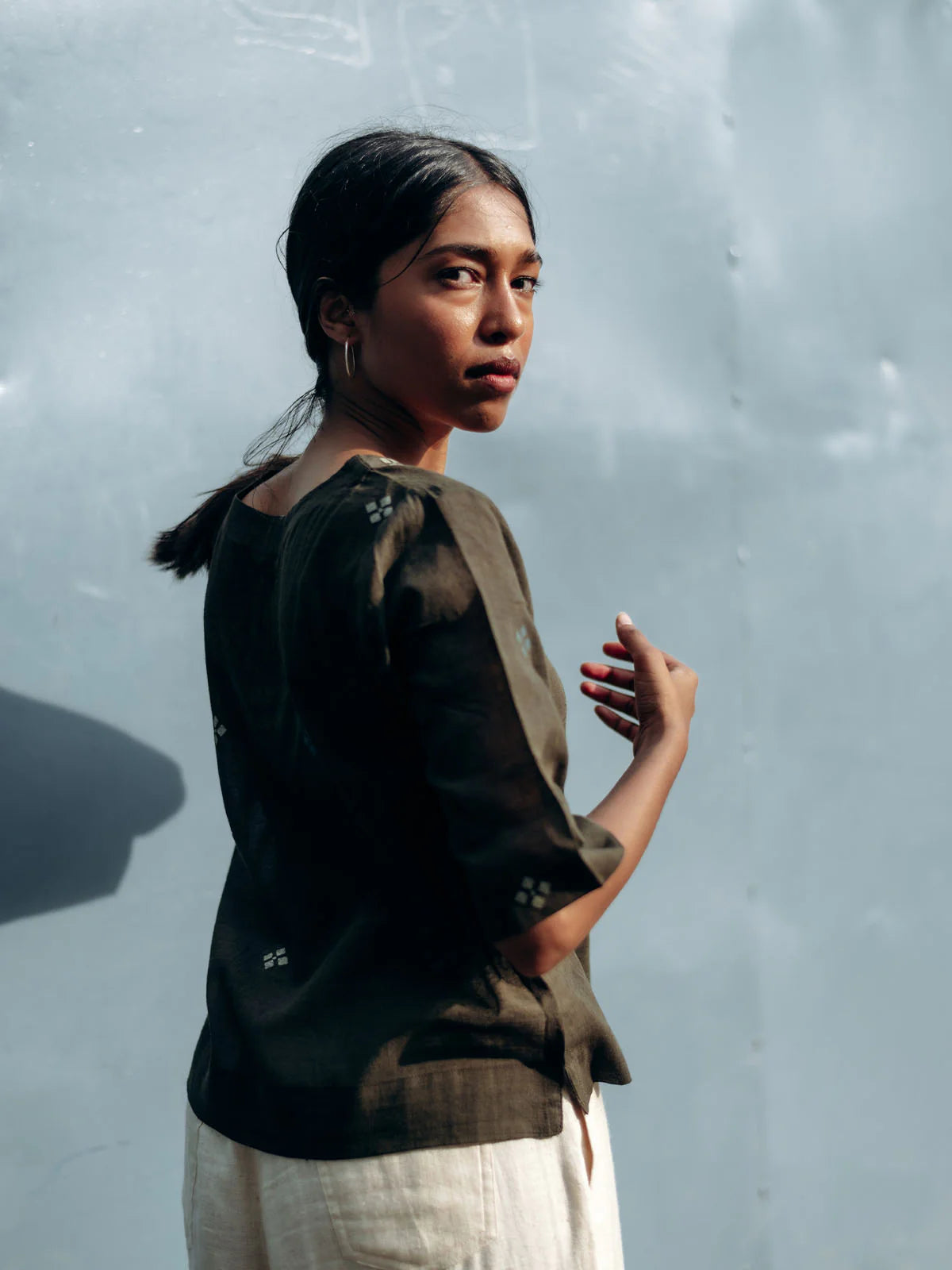 Woman wearing a gray top and light-colored pants against a textured gray wall.