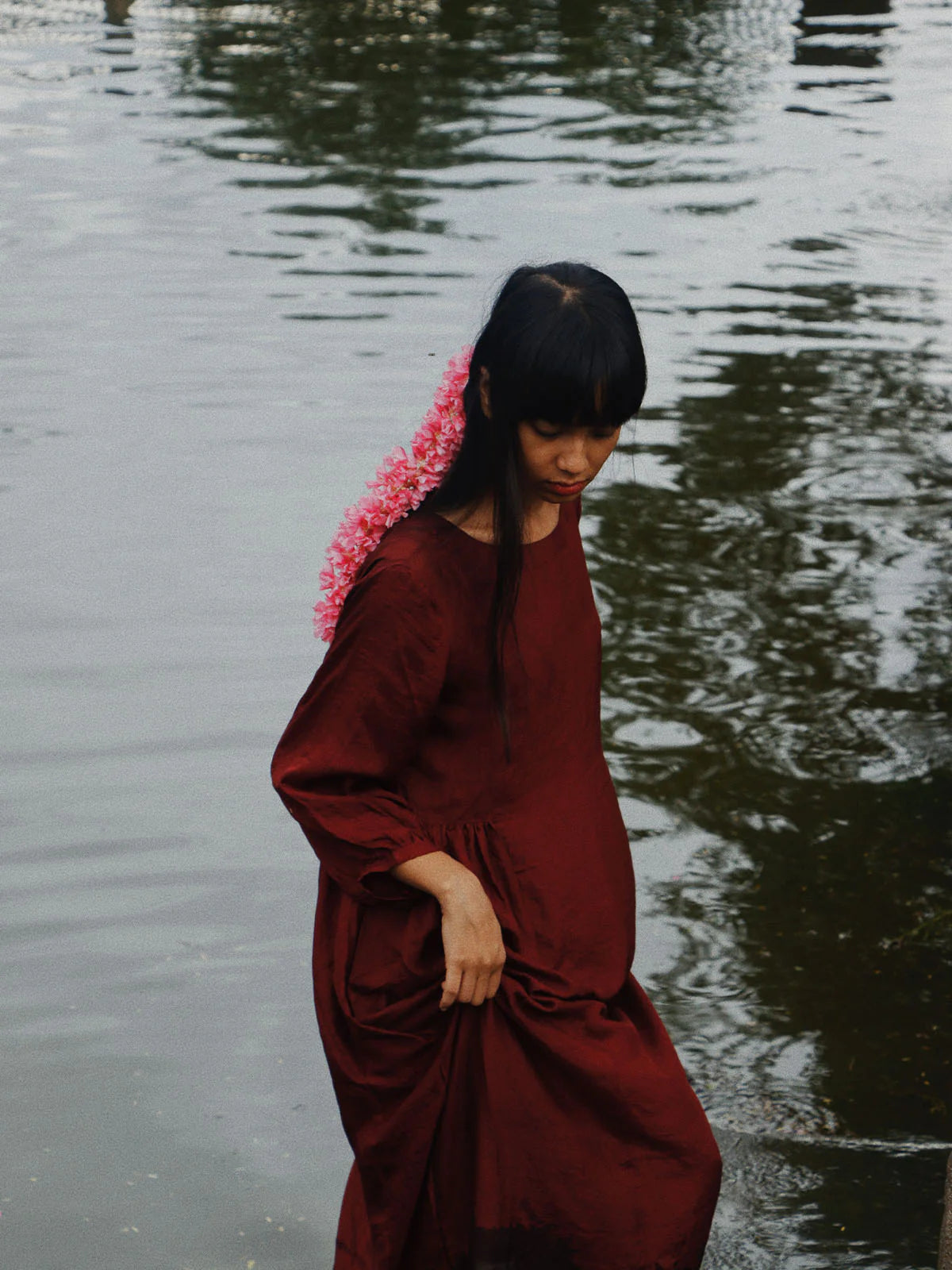 Woman in a red dress with pink flowers in her hair standing by a body of water.