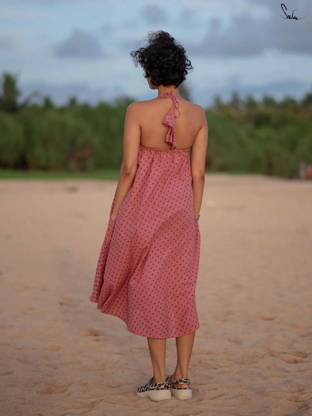 Woman in a pink dress standing on a sandy beach with greenery in the background