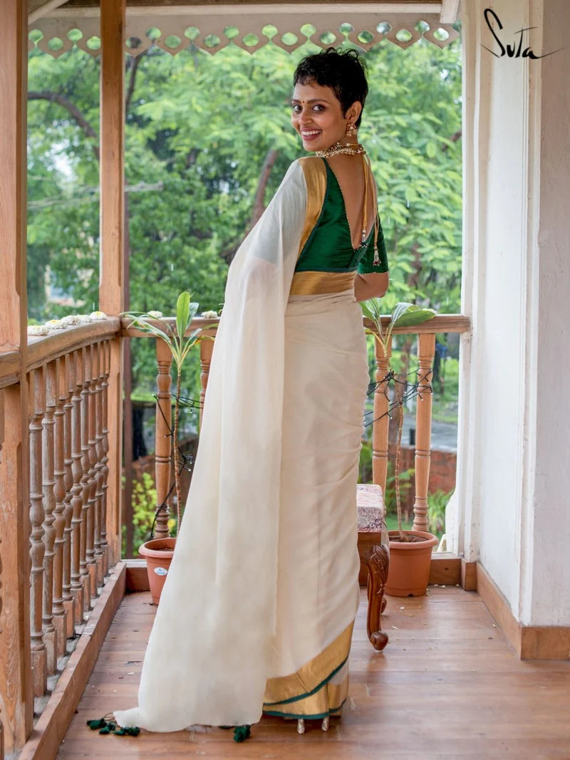 Woman in a green blouse and white saree standing on a wooden porch with greenery in the background