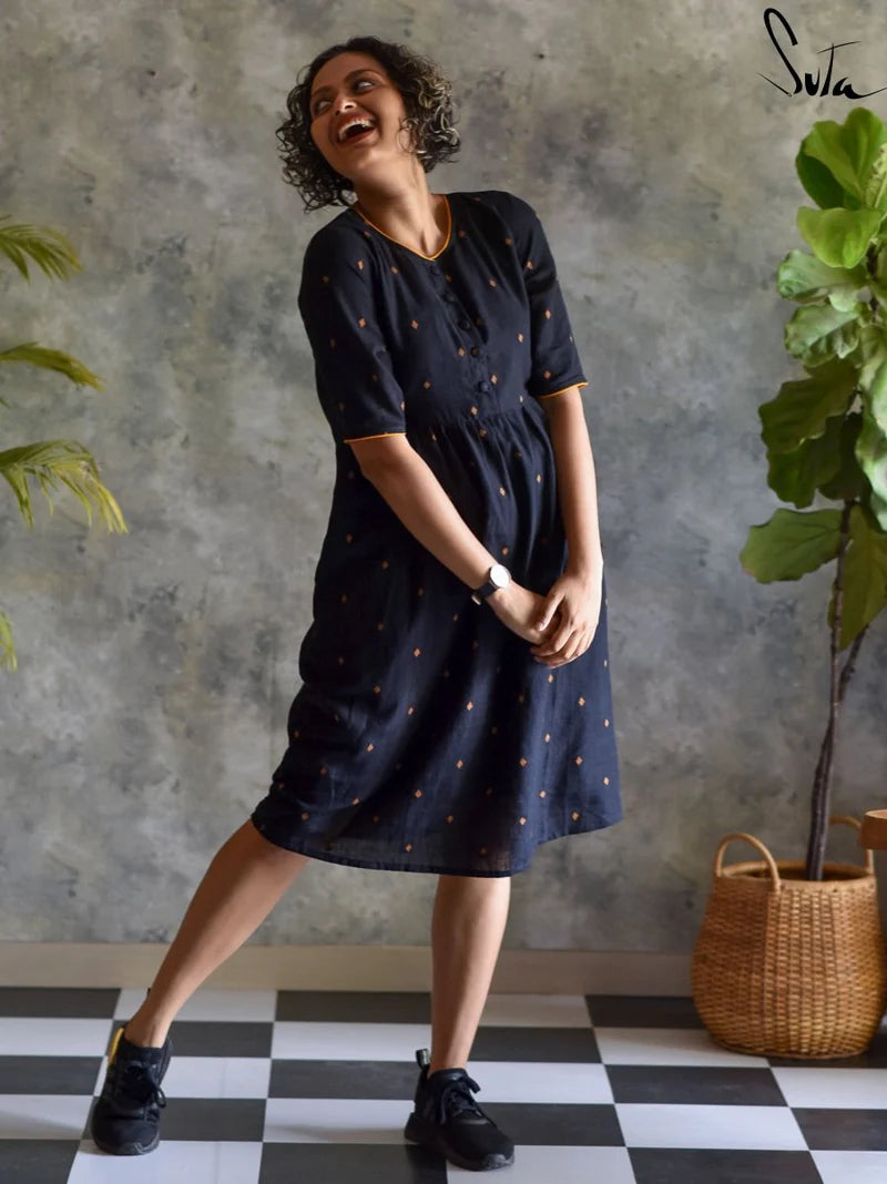 Woman wearing a black dress with gold patterns, standing in a room with a checkered floor and plants.