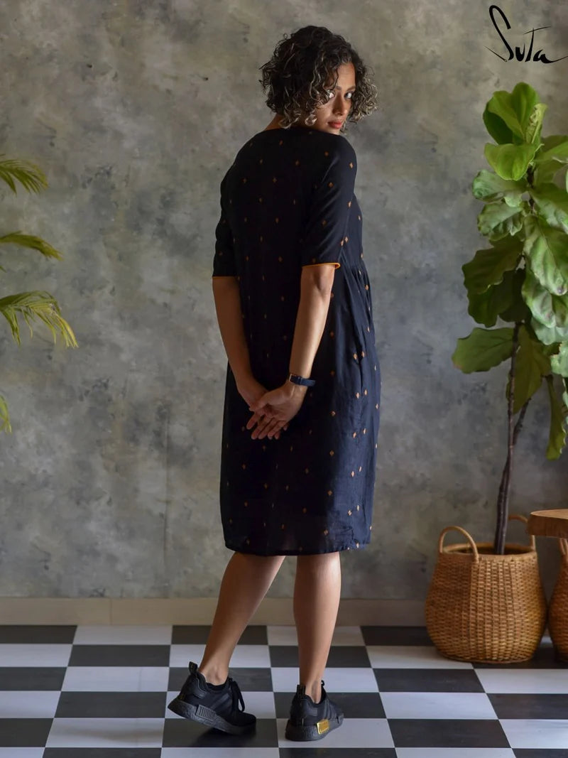 Woman wearing a black dress with gold patterns, standing in a room with a checkered floor and plants.