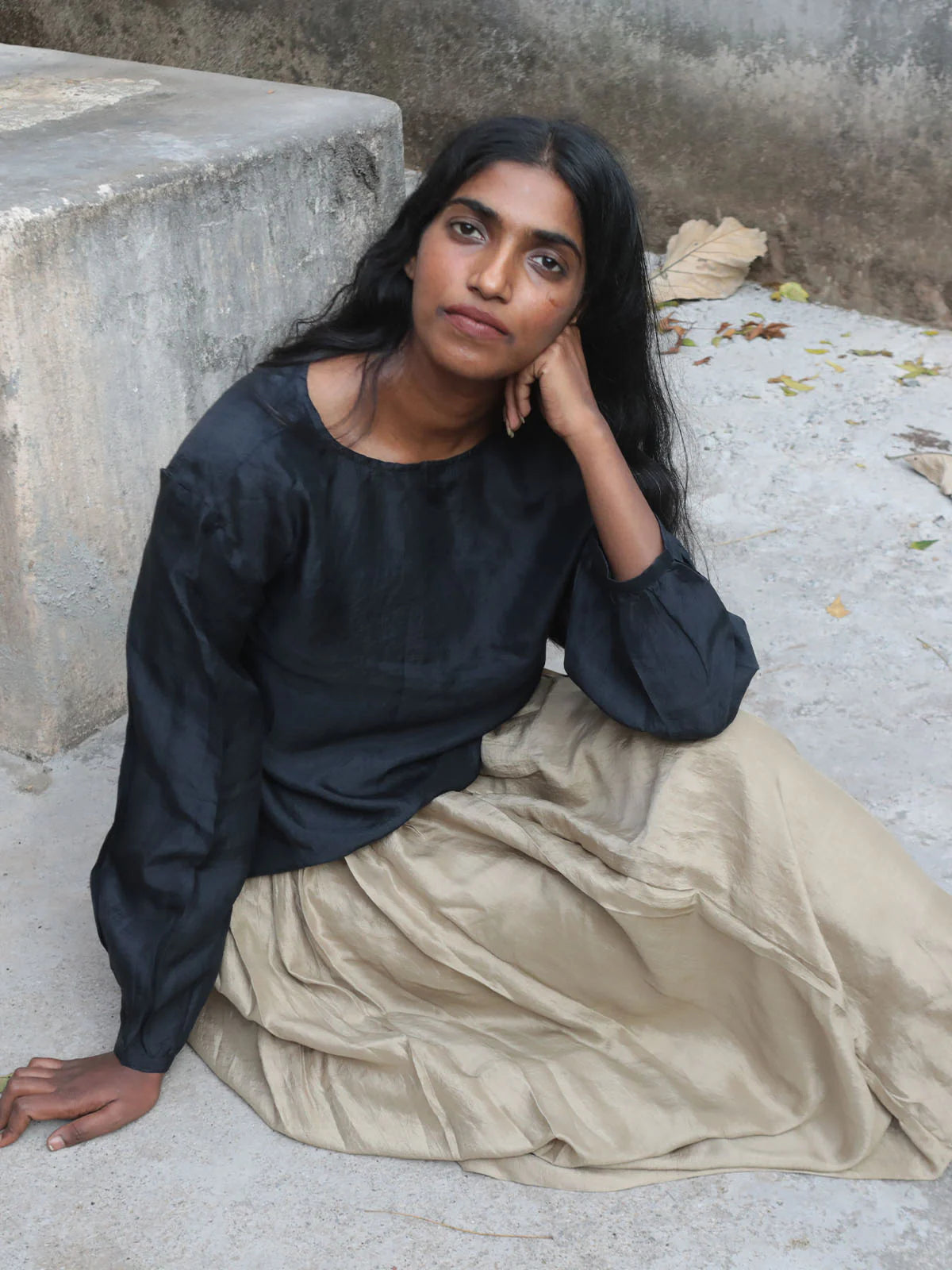 Woman sitting on a stone surface wearing a black top and beige skirt.