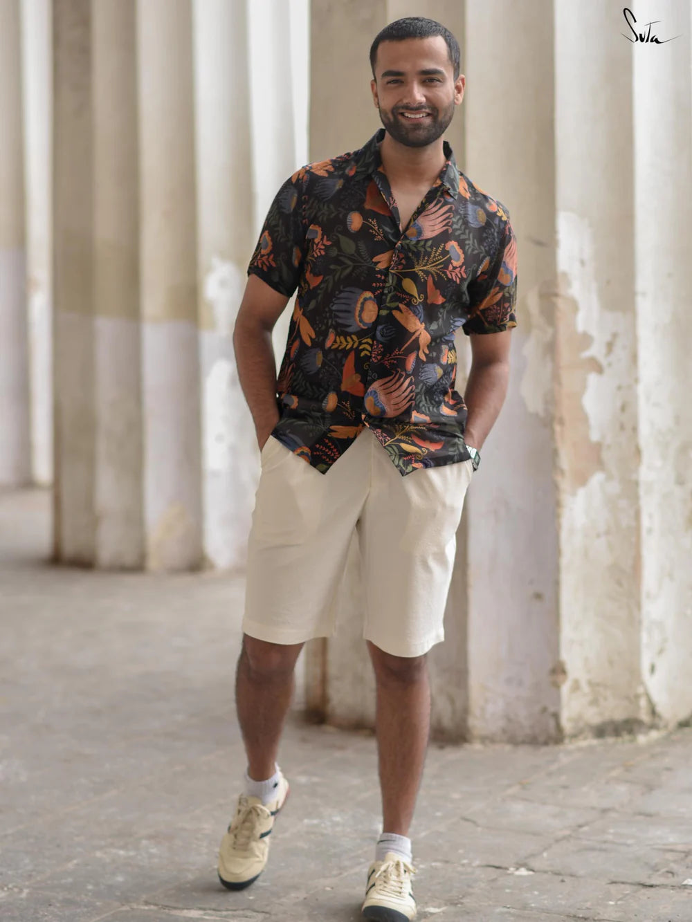 Man wearing a patterned shirt and beige shorts standing in front of columns.