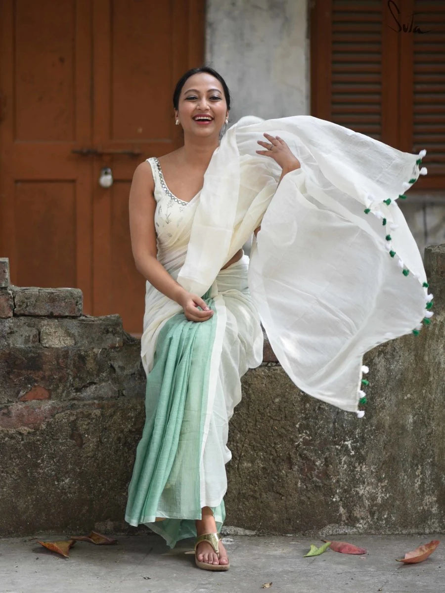 Woman in a white and green saree standing outdoors with a rustic background