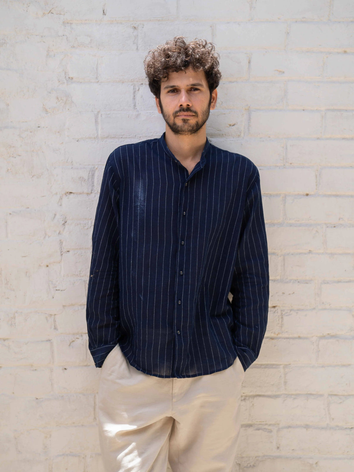 Man wearing a dark blue striped shirt and beige pants against a light brick wall.