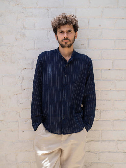 Man wearing a dark blue striped shirt and beige pants against a light brick wall.