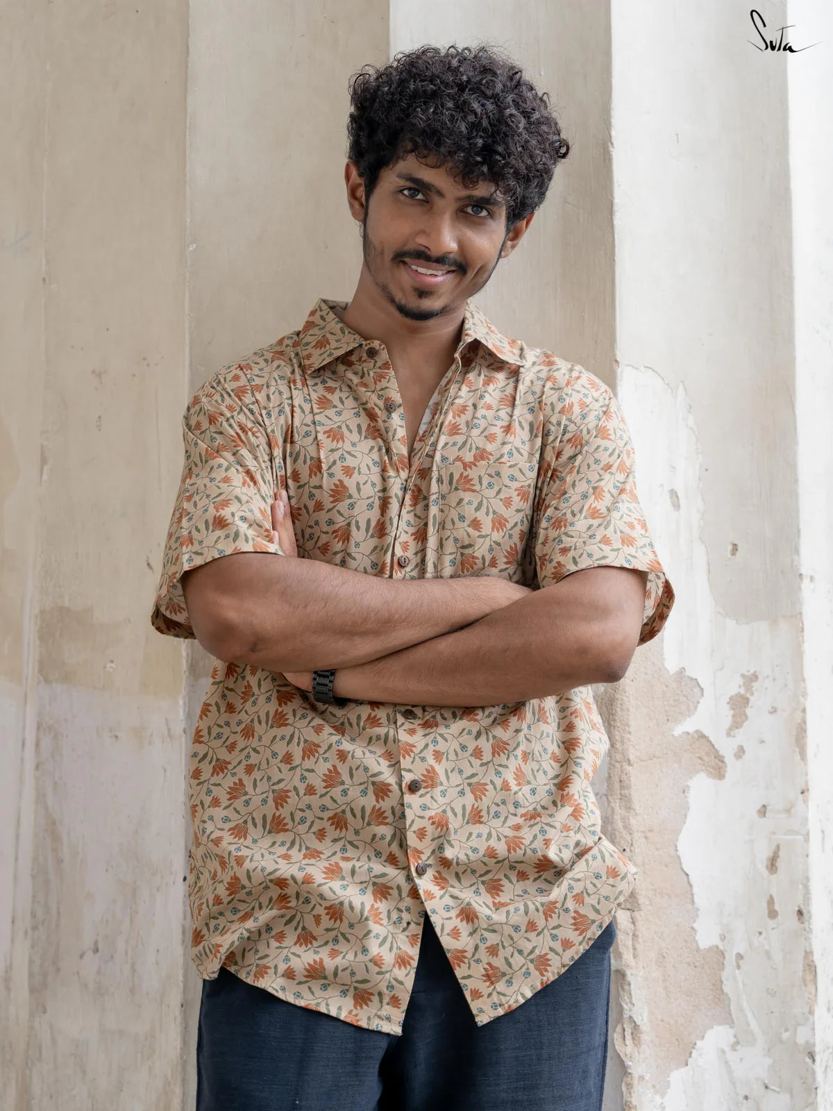 Man wearing a patterned shirt standing against a textured wall.