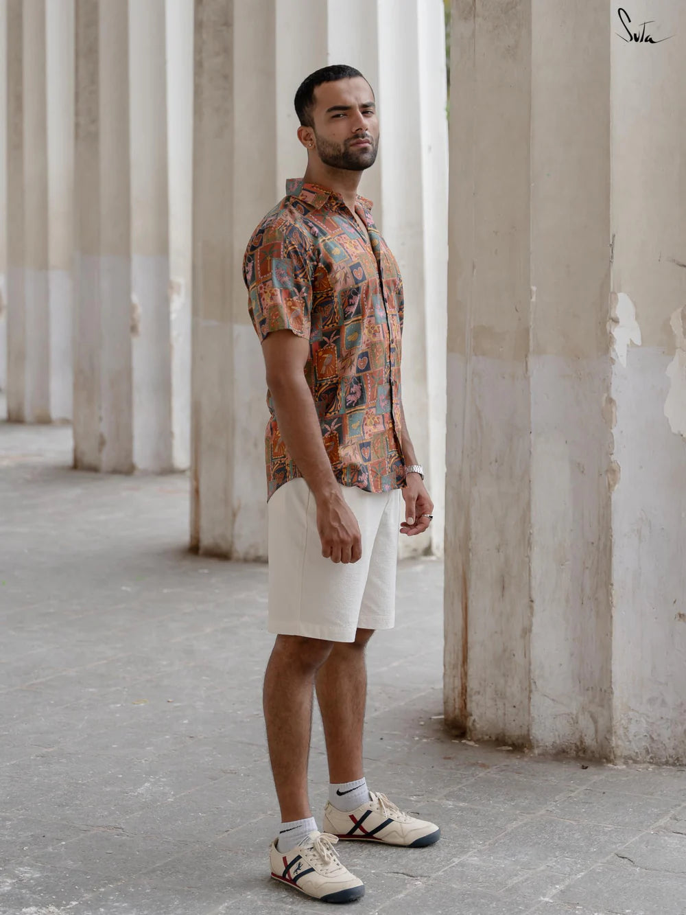 Man wearing a multicolored patterned shirt and beige shorts standing in an outdoor setting with columns.