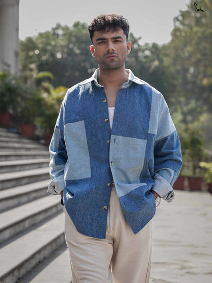 Man wearing a blue patchwork denim shirt standing on steps outdoors with greenery in the background