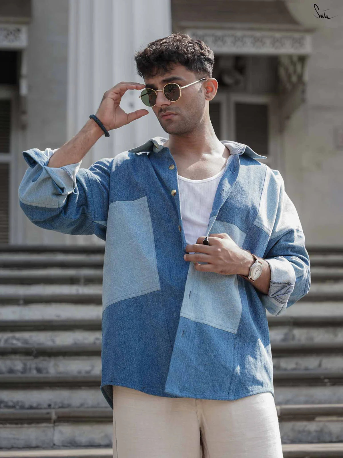 Man wearing a blue patterned shirt over a white shirt, holding sunglasses, with a building in the background.