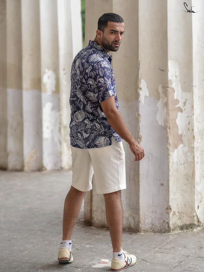 Man wearing a blue patterned shirt and white shorts standing in front of white columns.