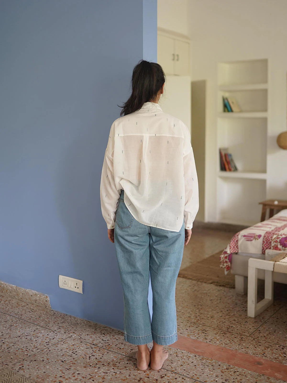 Person wearing a light-colored shirt and blue jeans standing in a room with a blue wall and a bed in the background.