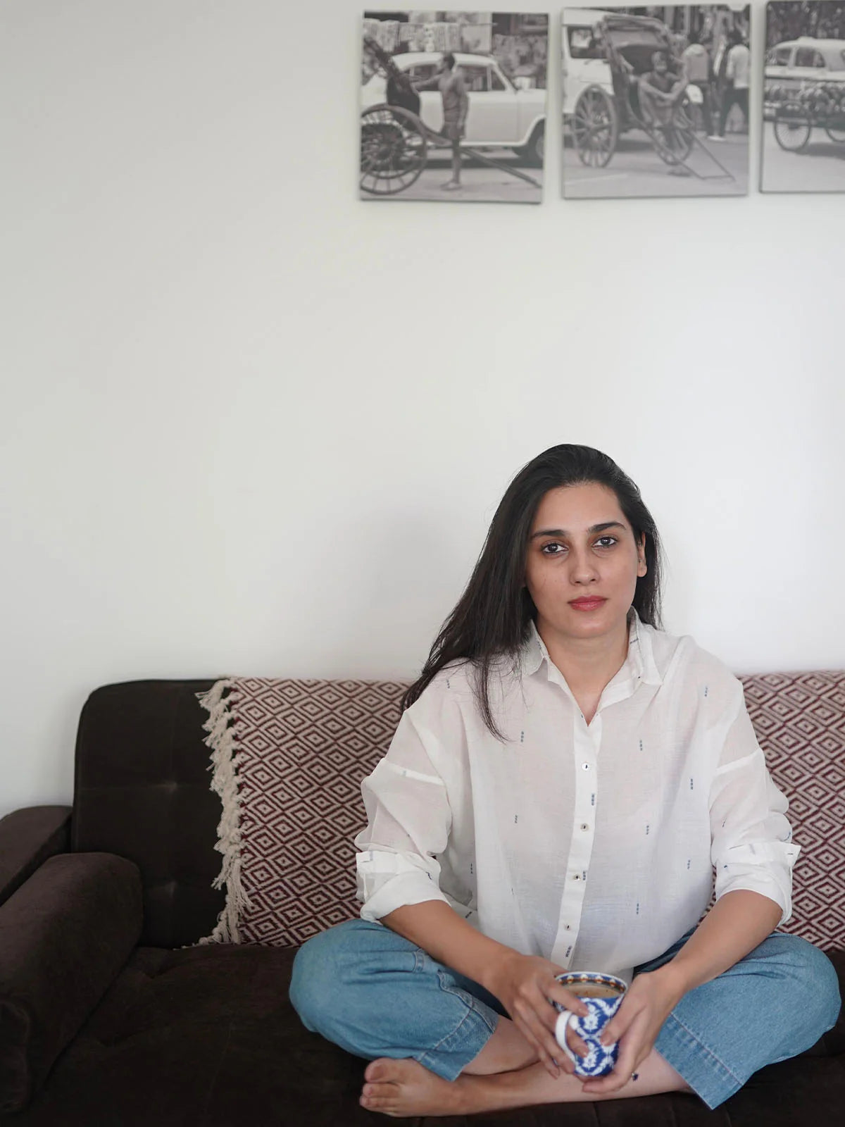 Woman wearing white shirt with small blue patterns sitting on a couch holding a cup in a room with framed pictures on the wall.