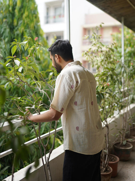 Person in beige shirt with half moon red pattern tending to plants on a balcony with buildings in the background
