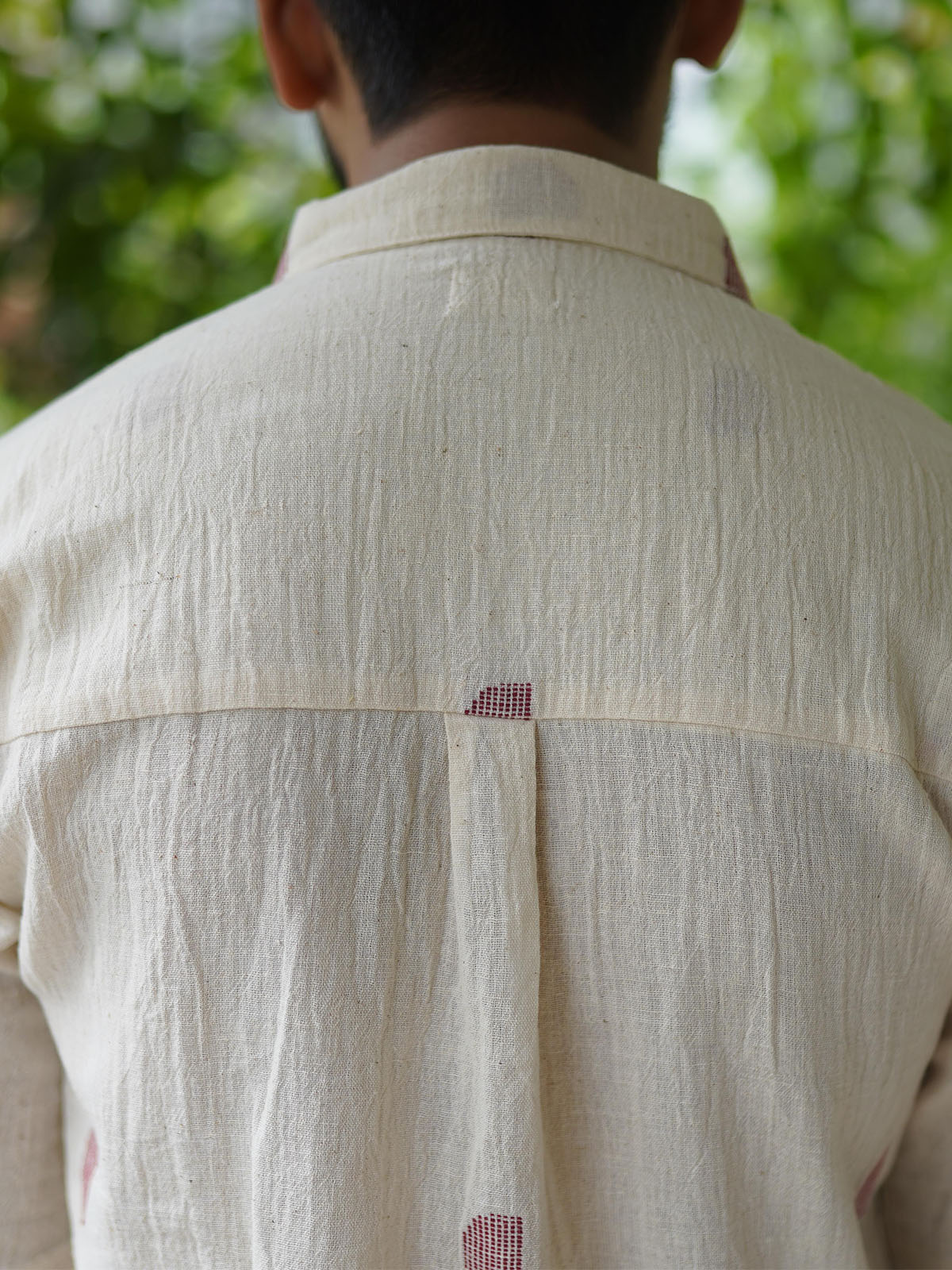 Person wearing a beige shirt with a subtle pattern, standing outdoors with greenery in the background.