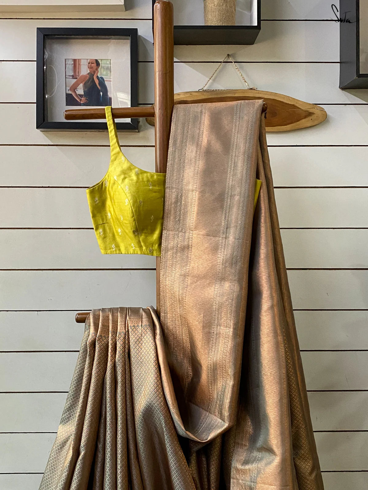 Gold saree draped over a wooden rod with a yellow top in the background on a wooden wall.
