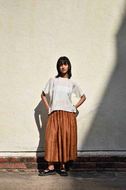 Woman wearing a white top and brown skirt standing against a light-colored wall.