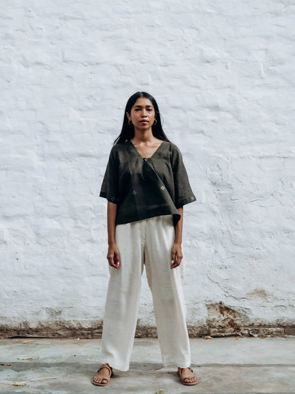 Woman wearing a gray top and white pants against a white brick wall.