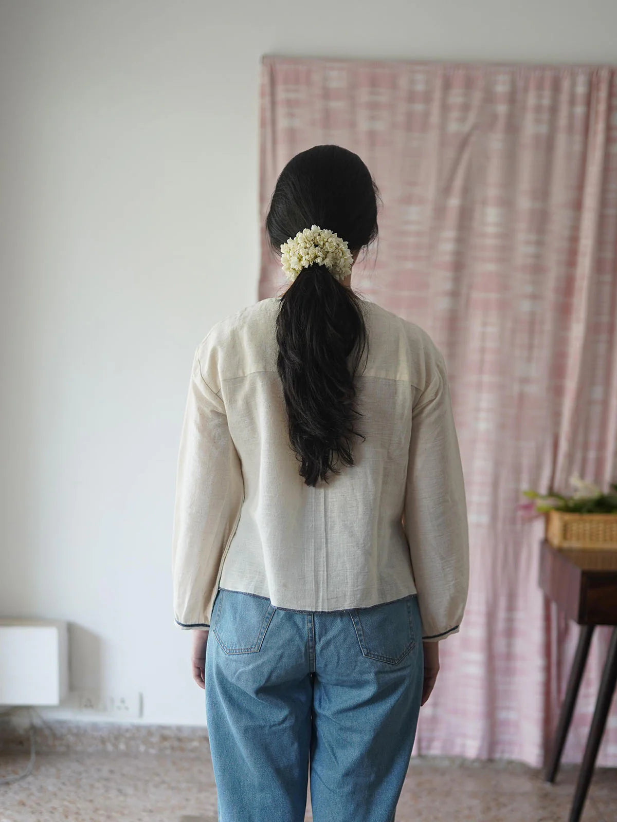 Person wearing a beige shirt and blue pants with a floral hair accessory, standing in a room with a pink tapestry.
