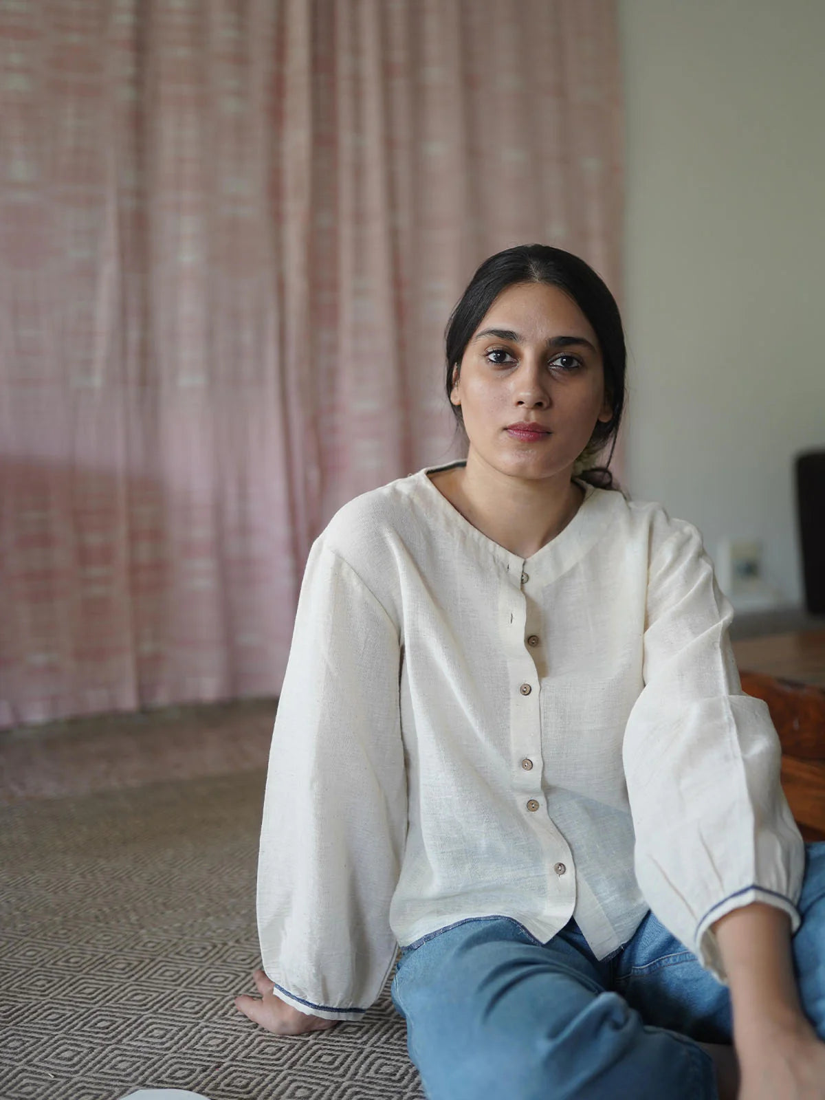 Woman sitting on a patterned rug in a beige shirt with a pink curtain in the background