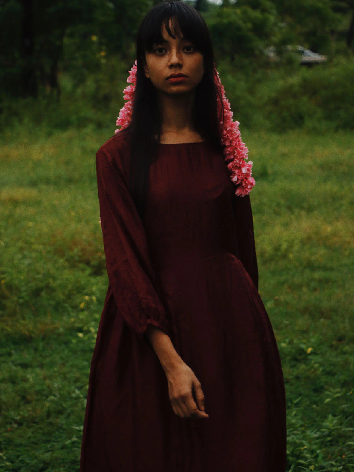 Woman in a dark red dress with pink floral headpiece standing in a grassy field.